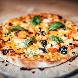 Eine Pizza mit fünf Basilikumblättern, Oliven und leckeren Käse liegt auf einem Holzbrett. Darunter sieht man das Holz eines Tisches.