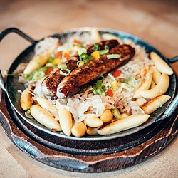 Zwei fränkische Bratwürste liegen wohl gebetet auf Schupfnudeln und Sauerkraut. Das Gericht ist in einem Eisenteller mit zwei Griffen angerichtet.