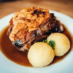 Auf einem weißen Teller sind zwei Kartoffelknödel mit Petersilie dazwischen angerichtet. Dahinter liegt das knusprige Schäuferl. Leckere Bratensoße zum Tunken füllt das weiße Teller aus.