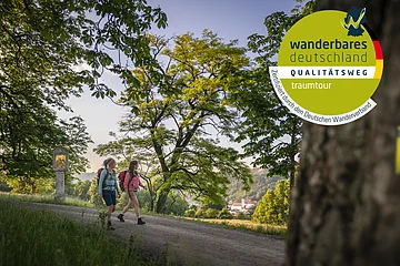 Zwei Wanderinnen auf einem Weg unter Bäumen, Wanderweg-Markierung an Stamm im Vordergrund
